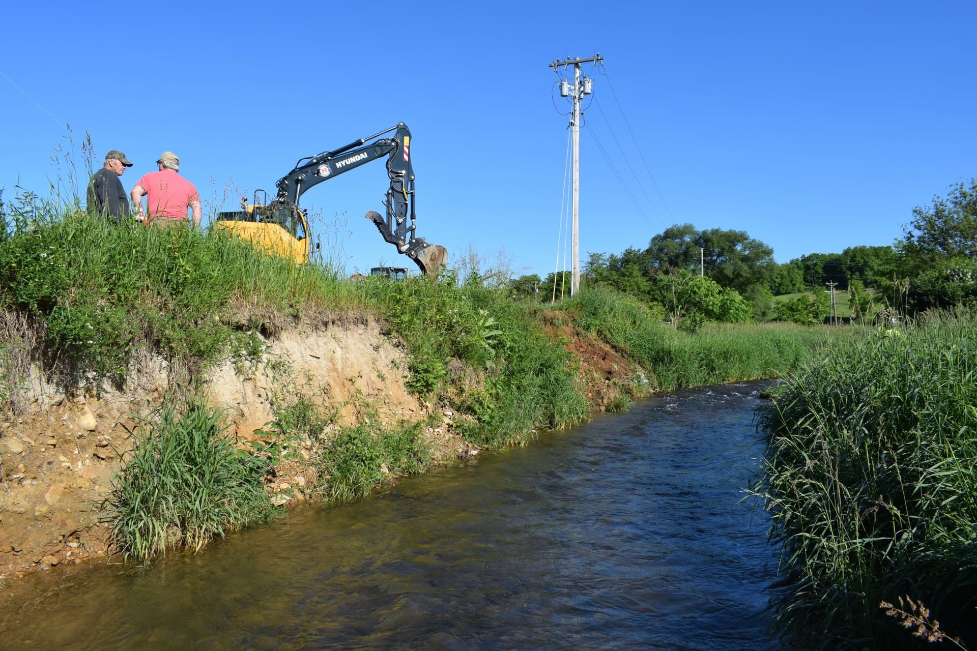 Hyundai excavator prepping the eroded streambank with two operators evaluating