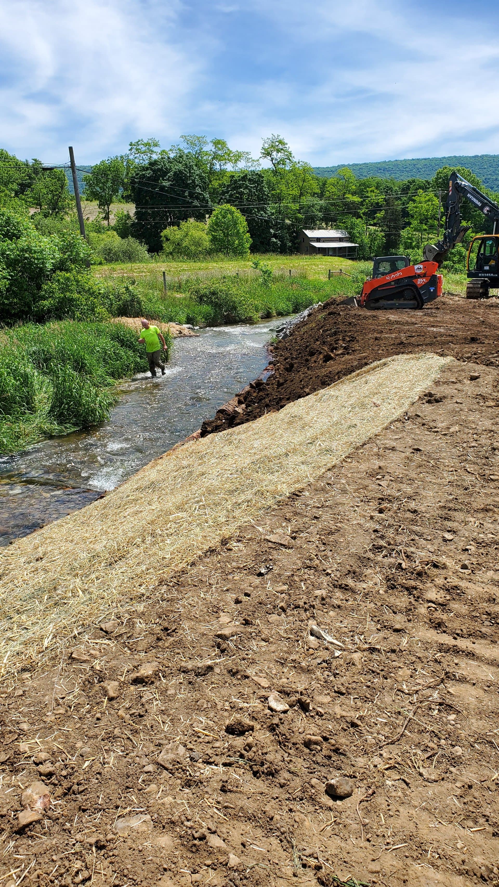 Skid steer beside freshly straw-mulched bank with mountains in the background